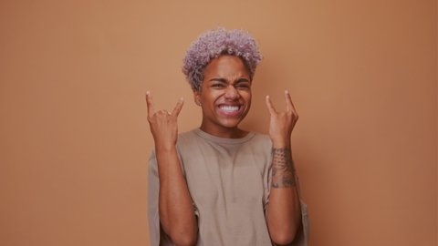 Positive joyful dark skinned woman makes heavy metal sign has fun on music rocking festival exclaims loudly closes eyes gestures actively, dressed in casual wear isolated over brown wall. Rock n roll.