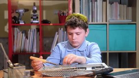 Schoolchild at desk with tools is intently repairing electronics. boy solders chip with soldering iron watches video with instructions in laptop. Online learning hobby. Creative development children