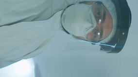 Vertical video: Microbiologist in ppe suit standing in laboratory looking at camera behind the glass wall in equipped lab. Doctor examining virus evolution using high tech and chemistry tools for