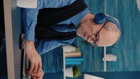 Vertical video: Focused old entrepreneur with headphones listening music while checking graphics writing on laptop working from home. Retired man using modern technology reading typing, searching on - Powered by Shutterstock - Get 15% off with code: PIKWIZARD15