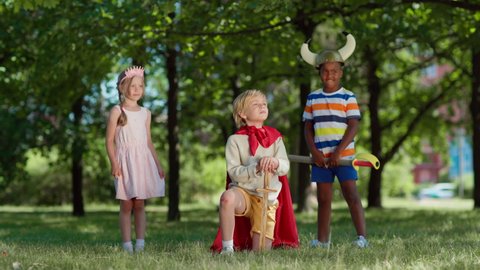 Diverse kids in costumes role playing in park. Little boy in red cape being coronated kneeing on grass with sword in his hand. Ecstatic boy raising arm with sword up. Children applauding to king