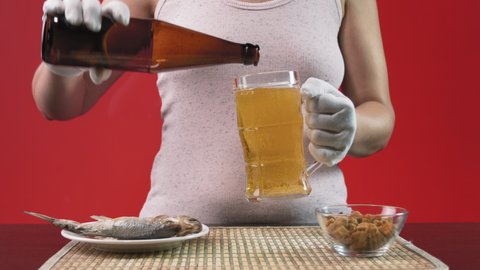 A girl bartender in white gloves pours beer from a bottle into a mug. There is a snack on the table next to it. These are two fish (roach) and crackers for beer. The girl stands on a red background.