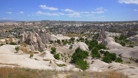 4K stock video footage of beautiful sunny summer landscape of Cappadocia. Rocky mountains, scenic valleys, green gardens of rural farms, small houses of Goreme city, blue sky and horizon line
