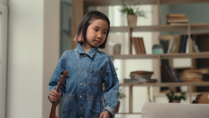 Little girl playing music on ukulele or small guitar. Happy asian kid laughing with smile on face and having fun time. Creativity in hobby of musical child or home leisure in comfort of living room