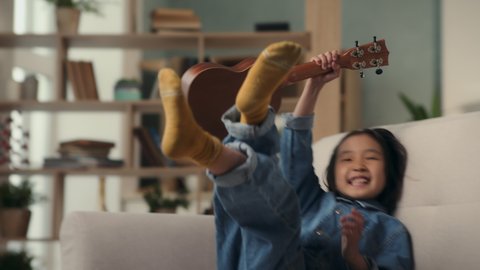 Little girl playing music on ukulele or small guitar. Happy asian kid laughing with smile on face and having fun time. Creativity in hobby of musical child or home leisure in comfort of living room