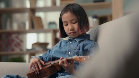 Little girl playing music on ukulele or small guitar. Home leisure or hobby of attentive asian kid. Happy musical child spending time in comfortable living room. Stringed instrument in hands closeup