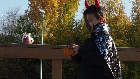 boy with black makeup for halloween, zombie. Scary little boy smiling wearing skull makeup for halloween halloween pumpkin lantern, sweets, with devil's horns and a cloak