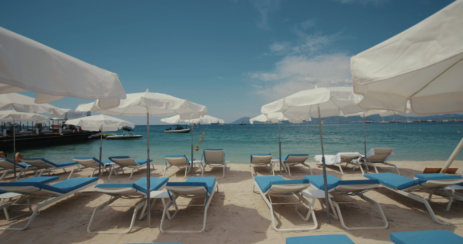 Sandy beach with deckchairs and parasols. Traveling forward towards the sea with yachts and mountains on the horizon, good weather and heat on vacation. Cannes film festival | Shutterstock HD Video #1075962398