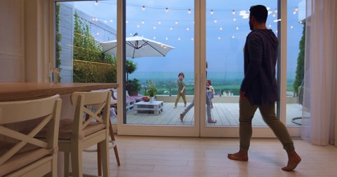 happy man opens sliding doors, going out on rooftop patio with playing kids and beautiful landscape view