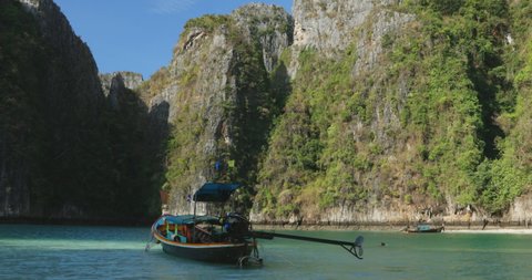 Static shot of longtail boat anchored in front of the famous karst island of Koh Phi Phi, Phuket Thailand. Boat rocks in the blue lagoon of Pileh Bay. Sun shines on cliff and tree background.