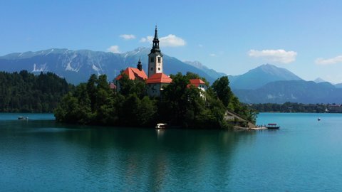 Pilgrimage Church of the Assumption of Mary In Lake Bled. Bled Island In Slovenia With Castle Museum On Precipice And Rocky Mountain In Background. orbiting shot