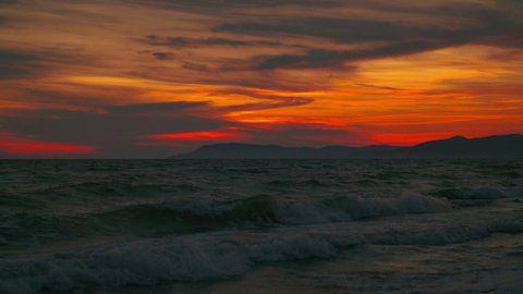 4K UHD Cinemagraph seamless video loop of of the sunset seen from a romantic sandy beach with mountains and waves at the beautiful Italian Mediterranean seaside with red and orange clouds in the sky.