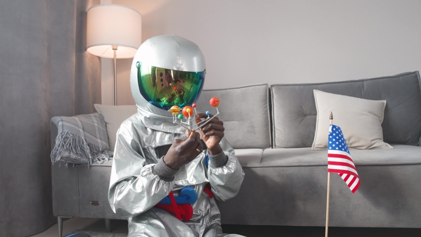 African man in an astronaut costume sitting on the floor in living room at home and playing with a toy model of the solar system, study of astronomy, man on self-isolation, 4k slow motion.