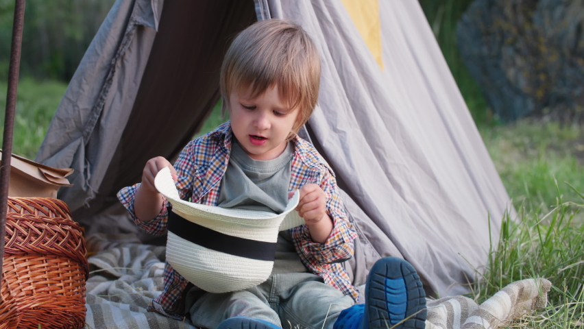 little cute male child playing with a hedgehog in a hat while sitting in a wigwam on a green lawn on a warm summer evening