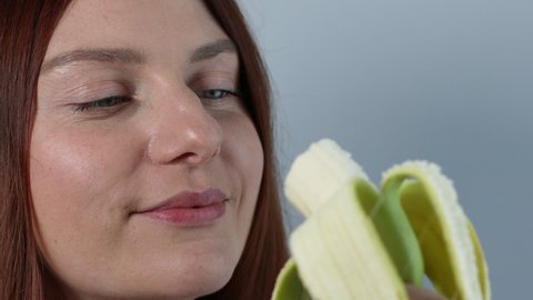 Close up of young caucasian beautiful woman biting yellow ripe banana and looking into camera. Face eating banana on grey background.