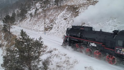 Siberia, Russia- january 22 2020: aerial steam locomotive train engine rides through picturesque winter heavy snow blizzard nature, releases steam clouds of smoke from chimney railway railroad. Baikal
