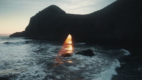 Cinematic golden sun beam coming through natural arch or hole in rock or island. Sunset ray reflecting on water surface with glow and glare illuminating sea water. Scenic seascape nature background 4K