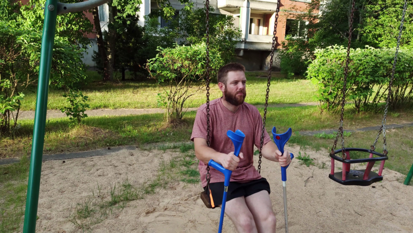 Bearded man with broken leg sitting on swings In playground and enjoying the summer weather. Close up of legs of adult man with crutches, because of broken heel.
