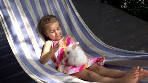 A girl plays with a white fluffy rabbit on a hammock. The kid communicates with her pet. Rabbit in the family. An unusual pet and a little girl. The concept of friendship between a child and a pet.
