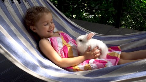 Happy little girl with positive emotions palys with her pet.A white fluffy bunny in the arms of a cute girl. The child holds a decorative rabbit in his arms and rides on a hammock on a sunny day. 