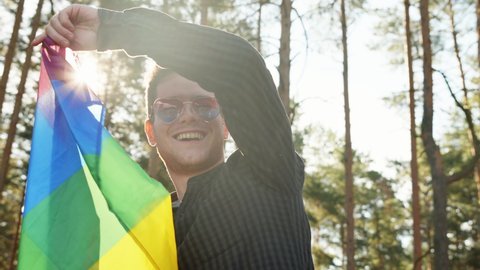 Homosexual man waving rainbow gay flag on sun flare light in beautiful summer day. Happy guy wearing heart sunglasses demonstrate his rights. LGBTQI, Pride Event, LGBT Pride Month, Gay Pride Symbol