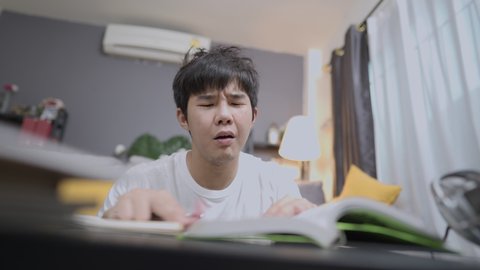 Unhappy Asian young student having a hard time study abroad, self study at home, unhappy student upset with homework on table in living room, problems in college, failure and success university life