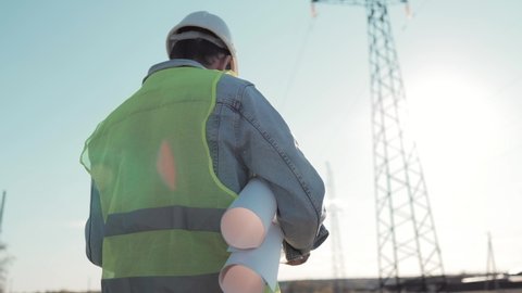 Architect Worker Checking Construction Project On Electric Tower. One Engineer Stands Near Power Lines At Sunset. Male Technician Is Operating A Documents Beside Electrical Transmission Lines.