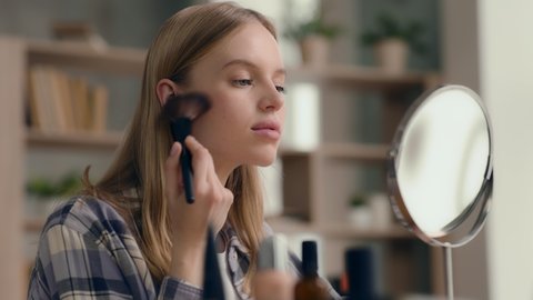 Young woman applying everyday makeup and preparing go out. Nude make-up of style girl or daily care for clear face of cute 20s lady. Morning visage of casual model looking at mirror at home close-up