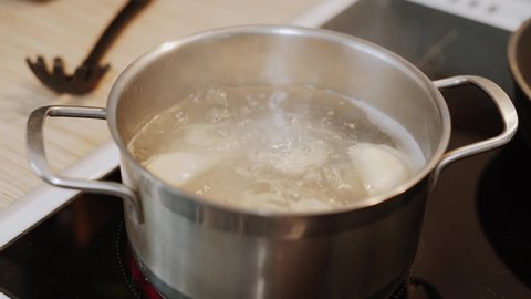 Water is boiling in stainless saucepan. Dumplings are cooked on electric home stove in the kitchen. Black spoon lies on the countertop.