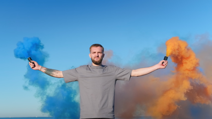 Young teenager with beard stands against background of blue sky and ocean holding firework colored smoke, colored bombs looks at camera. Orange and blue smoke bombs in the hands of man, man protests.