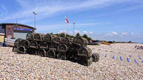 Selsey, West Sussex, UK, July 09, 2021. Stacked Lobster Pots and fishing gear on East Beach near the Selsey Lifeboat Station with the RNLI flag fluttering in the wind.