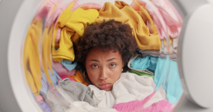 Sad Afro American woman poses in washing machine drum with clothes for washing falling on her has much housework feels tired loads washer with laundry. Frustrated housekeeper does laundering | Shutterstock HD Video #1076015747