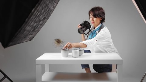 Inspired woman in casual clothes using digital camera for taking pictures of ceramic cups and candles at photo studio. Creative process of female photographer. Favorite hobby.