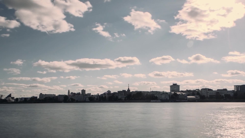 Summer landscape of white clouds flowing above the city and a wide river, timelapse effect. Video. Town located along the river on a windy day.