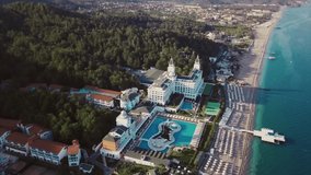 Aerial view of a luxury resort located by the sea. Video. Flying over the breathtaking hotel surrounding territory with the sea coast, swimming pool, pier, and green forest.