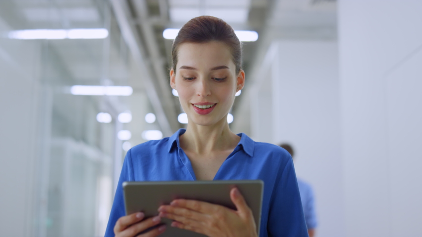 Beautiful businesswoman looking tablet screen while walking on modern office. Happy woman reading message on digital device indoors. Joyful business lady working while going along creative space.