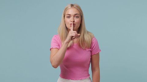 Secret smiling fun charming young blonde woman 20s wears pink t-shirt look aside say hush be quiet with finger on lips shhh gesture isolated on pastel plain light blue color background studio portrait