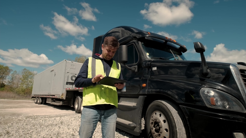 Truck driver standing by the truck in a yellow vest and using a tablet to fill a lookbook. Wide camera shot