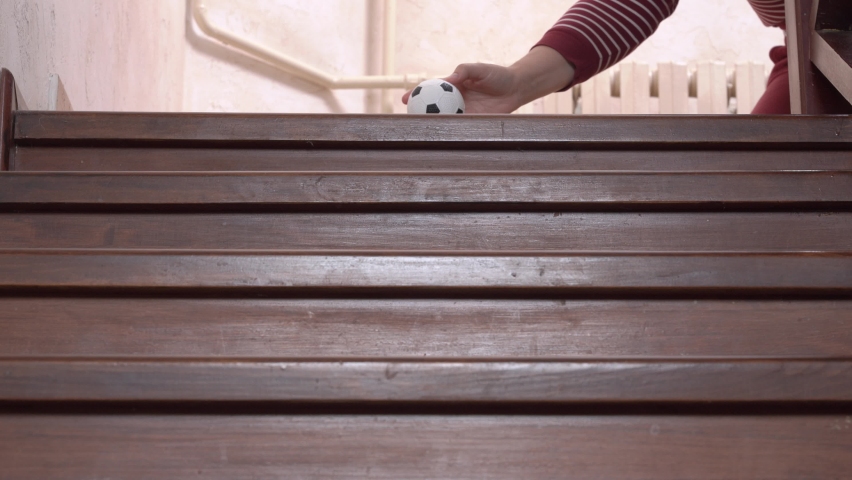 Person pushes smaller replica of soccer ball down the stairs, and toy jumps down wooden steps, close up, camera moves after the falling object, view from below.