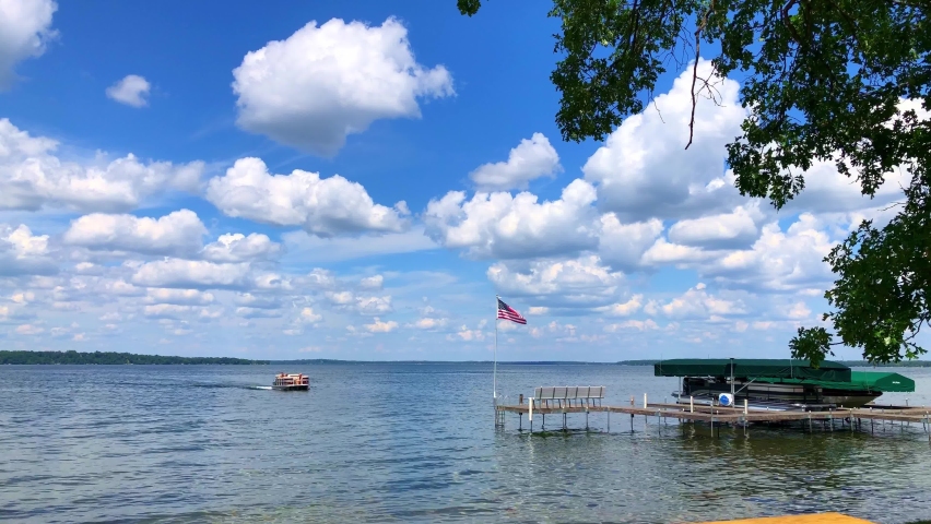 Beautiful Minnesota lake scene as a pontoon slowly approaches shore. A hand held clip with with an American flag waving over the boat dock, close tree branches, and fluffy white clouds under blue sky.