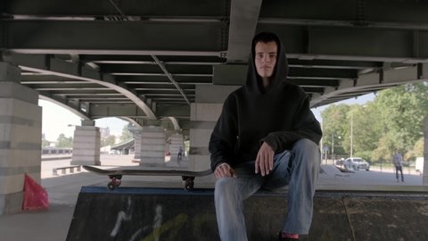 Slow motion video portrait young serious not smiling caucasian skateboarder, looking at camera. Guy is sitting on skateboard in black hoodie under an iron bridge on skate Park.