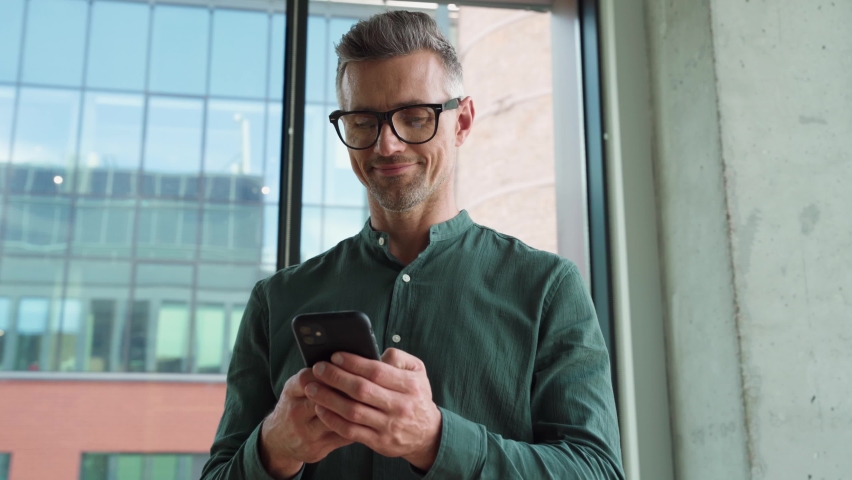 Smiling mature businessman holding smartphone standing in office. Middle aged manager ceo using cell phone mobile apps. Digital technology applications and solutions for business corporate development Royalty-Free Stock Footage #1076061548