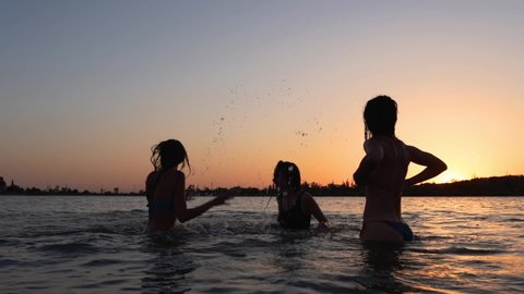 Happy wet girls silhouettes in bikini have fun splashing water to each other on sunset. Female friends have fun making splashes in pond on sunrise. Women go swim and bathe in lake in dusk. Slow motion