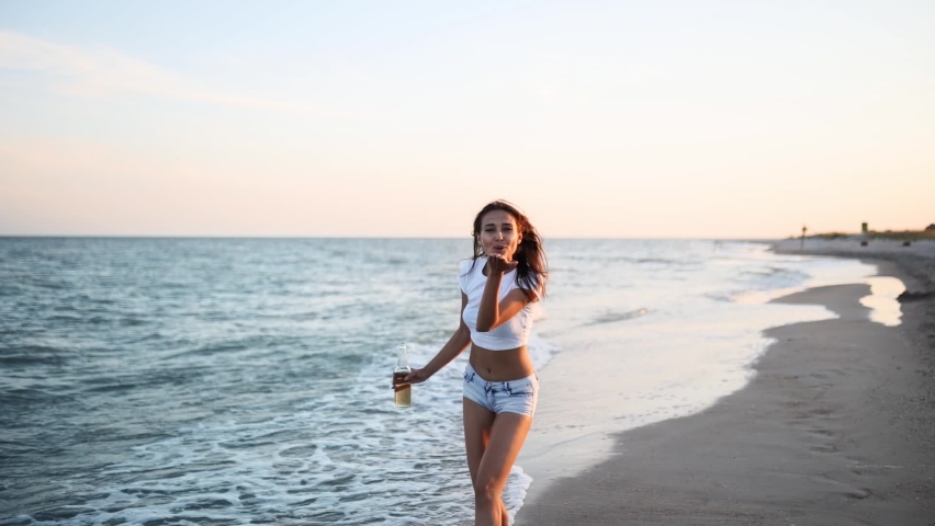 Pretty woman walking along seaside on a sandy beach having fun with beer bottle. Attractive fitted girl sends air kiss, enjoys summer vacation near sea waves with beverage. Follow me. Slow motion. | Shutterstock HD Video #1076066795