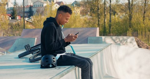 Dark-skinned teenager sits on concrete ramp, bike behind him, holds phone in hands, posts on social media, photo of bmx ride, brags to friends about new tricks