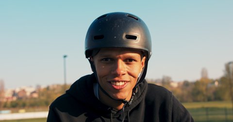 A teenager in a black sweatshirt and helmet stands on a ramp at a skatepark and smiles for the camera, his face illuminated by the setting sun