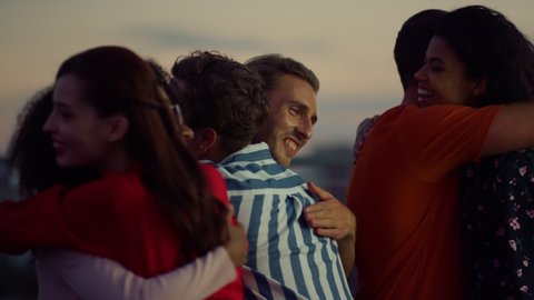 Cheerful friends greeting each other at rooftop evening party. Beautiful people hugging while meeting outdoors. Young buddies feeling happy together outside. Friendship concept.