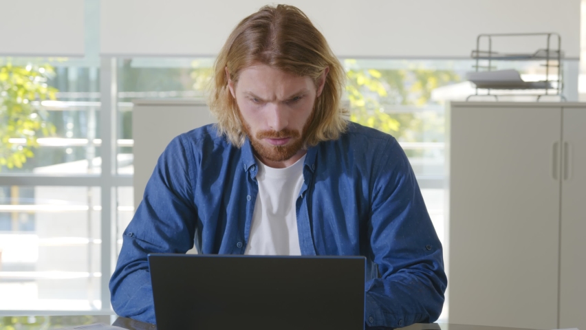 Tired young businessman in casual clothes feeling stressed and overwhelmed sitting in modern office with laptop. Portrait of frustrated bearded entrepreneur working on computer in office | Shutterstock HD Video #1076071835