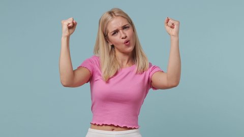 Excited jubilant overjoyed happy young blonde woman 20s wears pink t-shirt doing winner gesture celebrate clenching fists say yes isolated on pastel plain light blue color background studio portrait