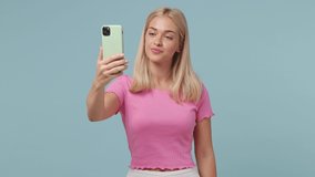 Smiling young blonde woman 20s wears pink t-shirt doing selfie shot on mobile phone post photo on social network blow send air kiss isolated on pastel plain light blue color background studio portrait